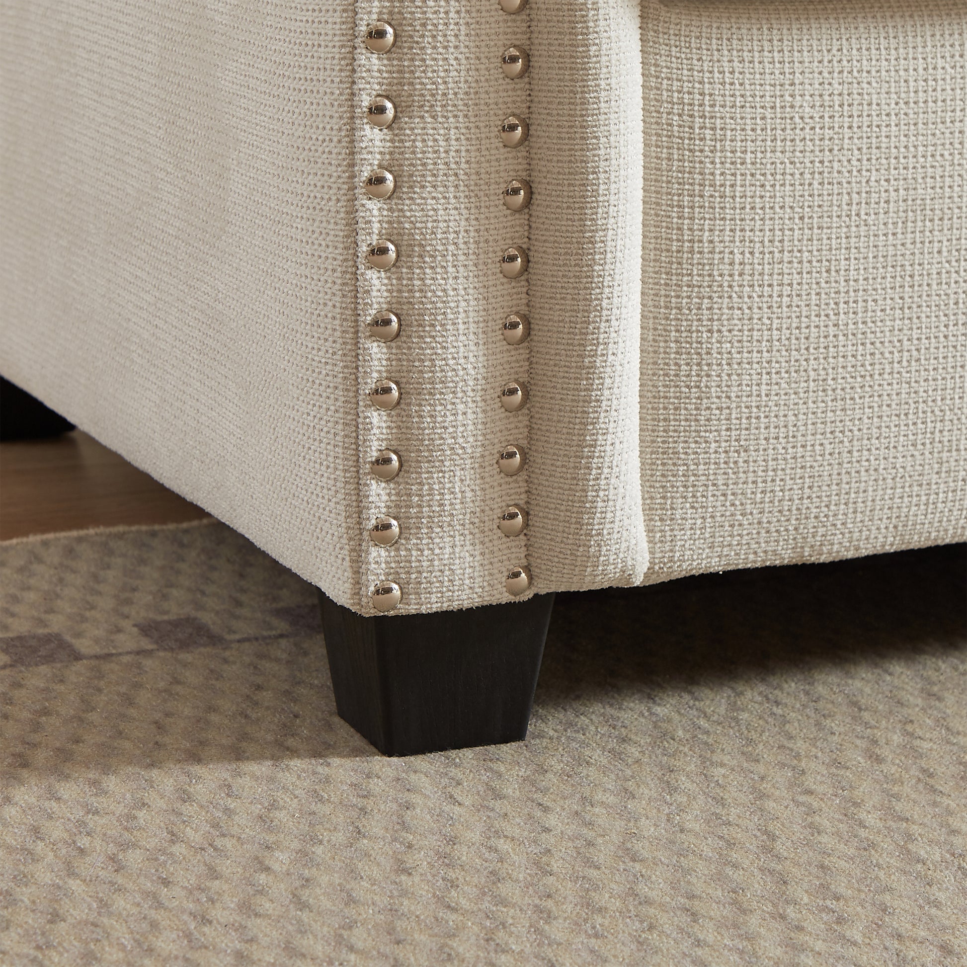 Textured beige armchair fabric with decorative silver nailhead trim and dark wood legs
