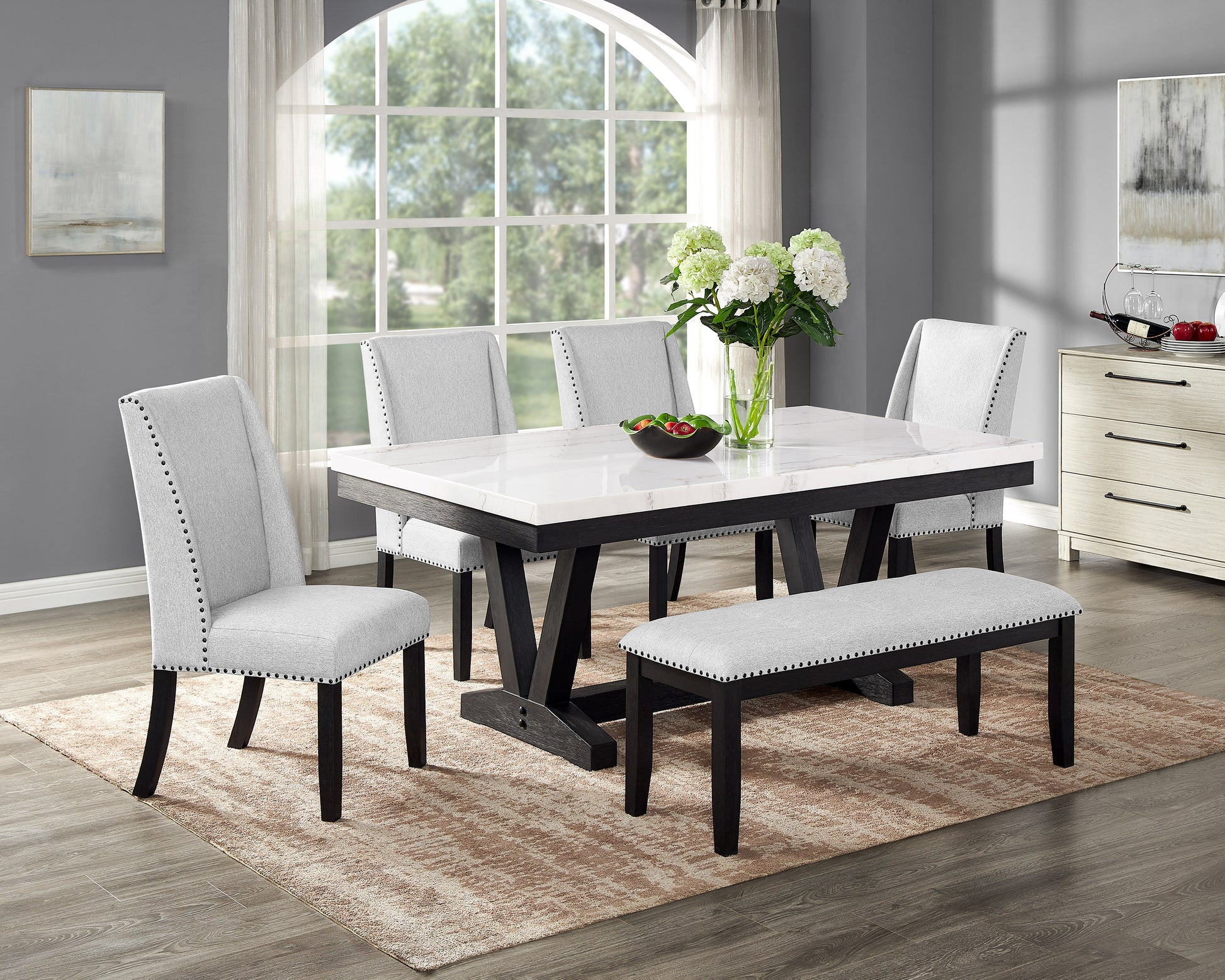 Gray upholstered dining chairs with nailhead trim surrounding a faux marble top dining table with dark wood legs