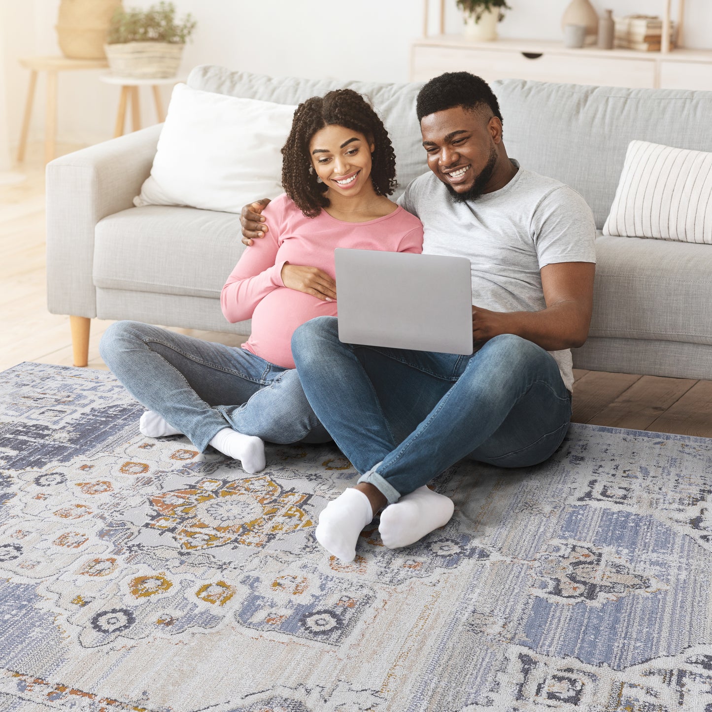 A pregnant couple smiles while looking at a laptop seated on a distressed vintage area rug
