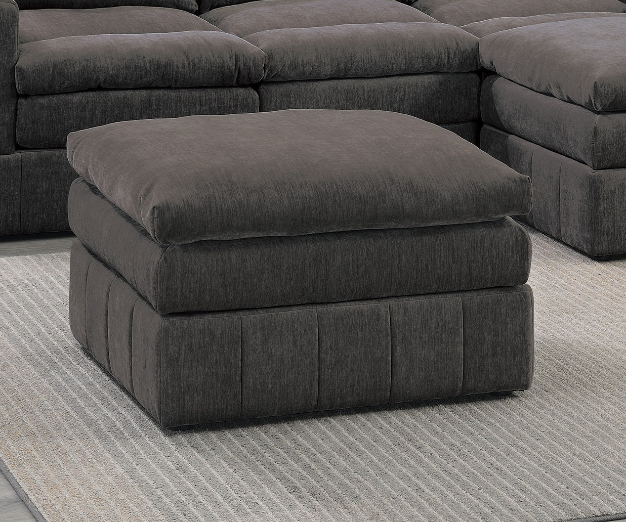 Deep charcoal grey upholstered ottoman with plush cushioning on a striped rug