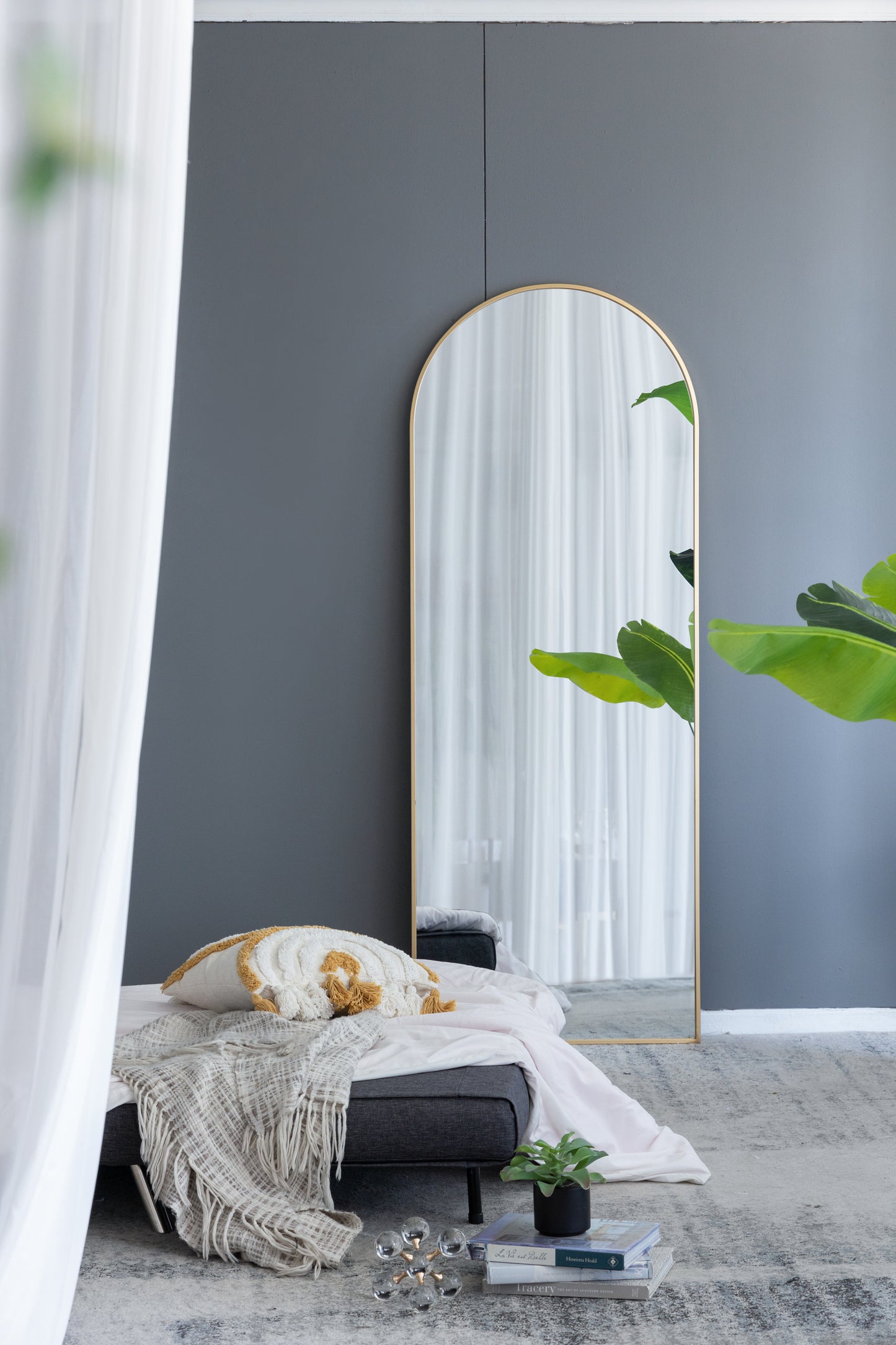 Tall arched floor mirror with thin gold metal frame reflecting white curtains