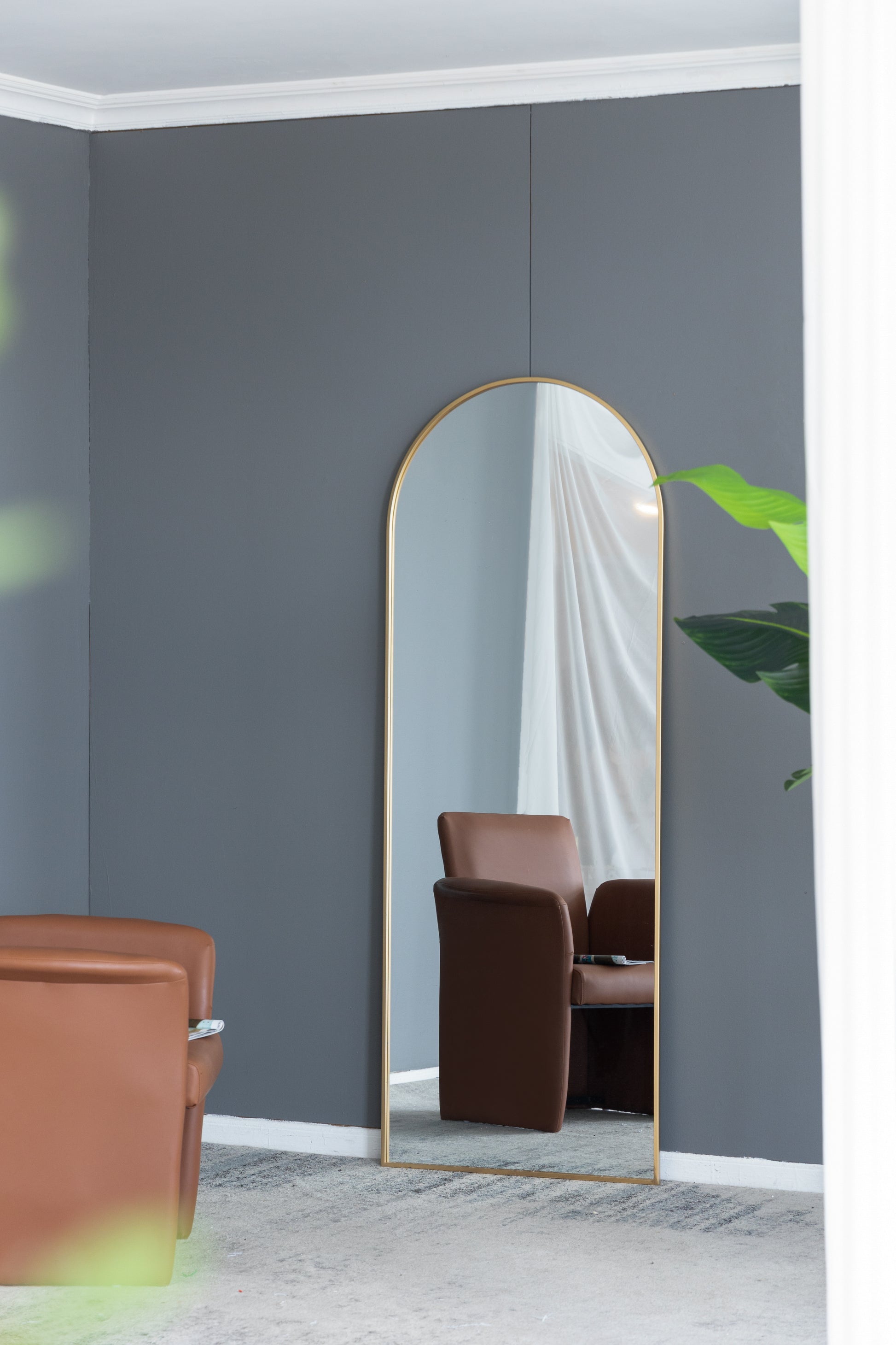 Tall arched mirror with gold frame reflecting brown leather armchair and white curtains