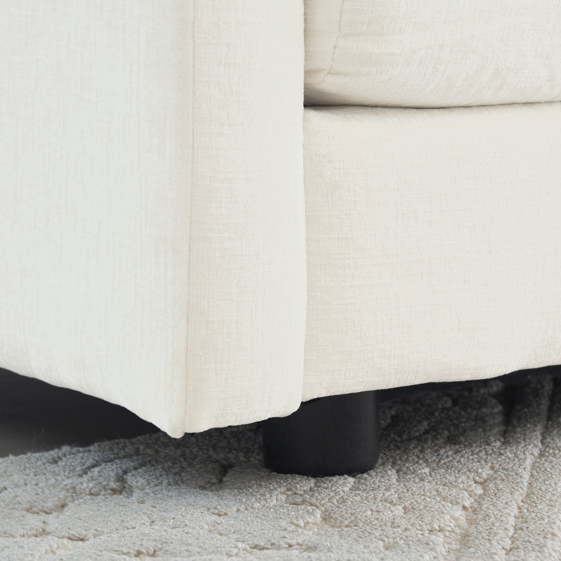 Close up of a cream textured fabric sofa arm with a black wooden leg resting on a plush rug