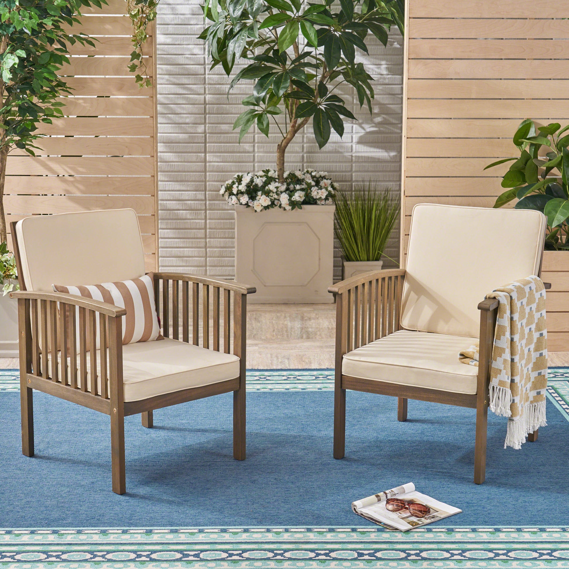 Two wooden patio chairs with cream cushions and striped pillows on a blue outdoor rug