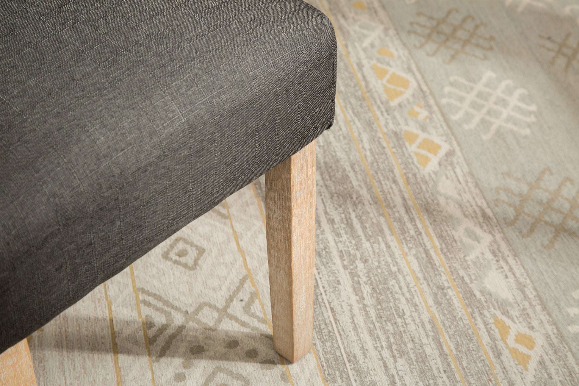 Closeup of a charcoal grey linen upholstered chair leg with natural wood finish resting on a patterned rug