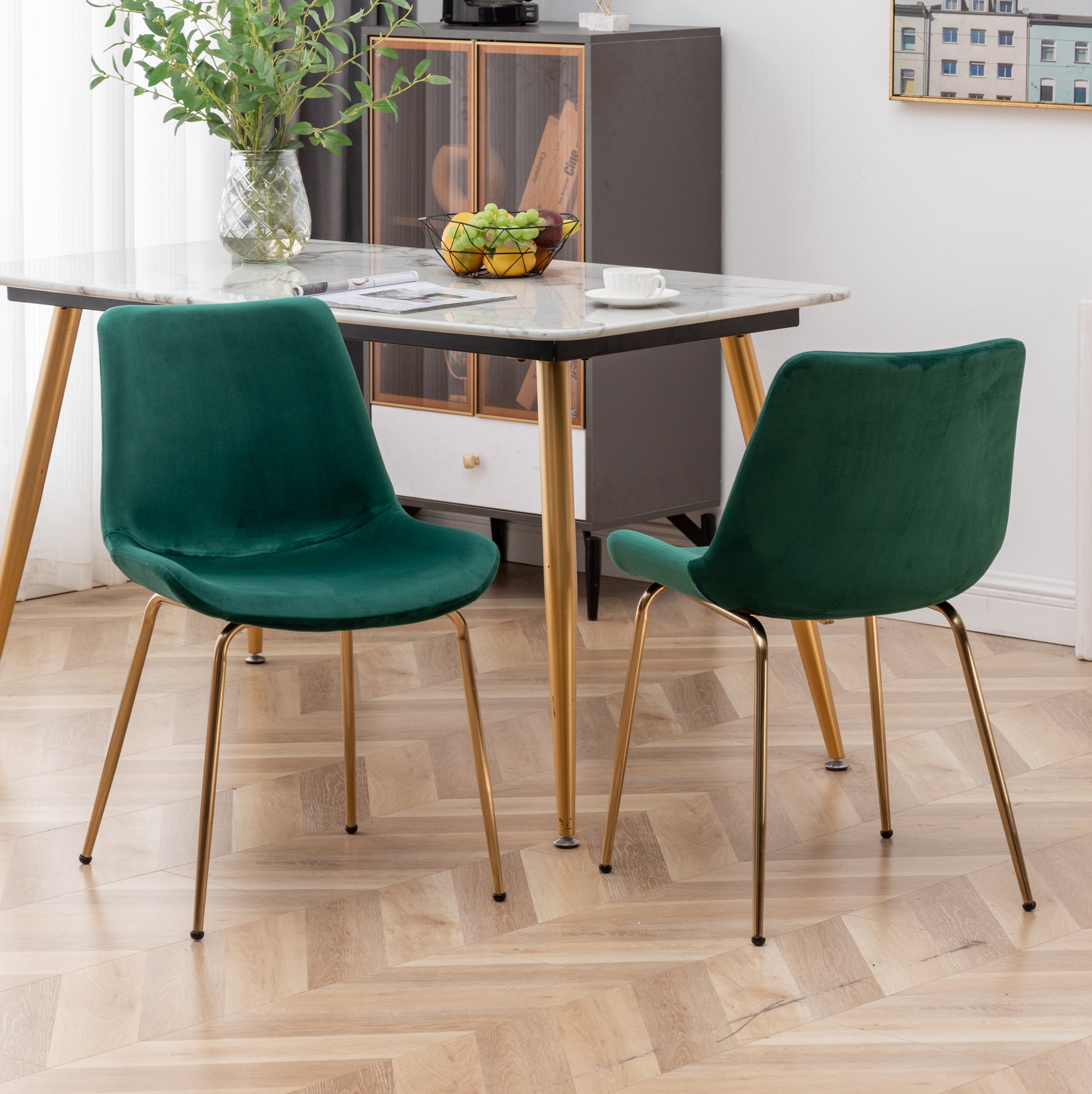 Two emerald green velvet dining chairs with golden legs arranged around a marbletopped table