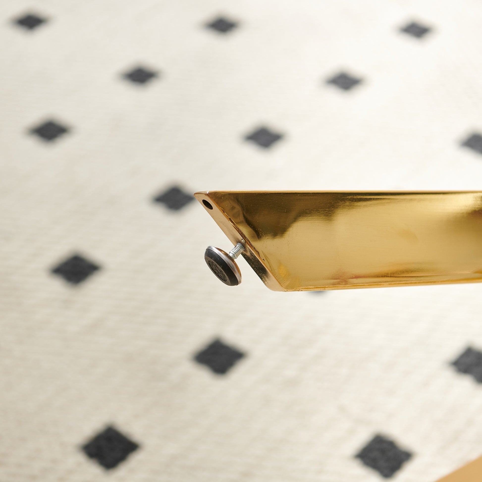 Glossy gold table leg with adjustable foot on a cream and black diamond patterned rug