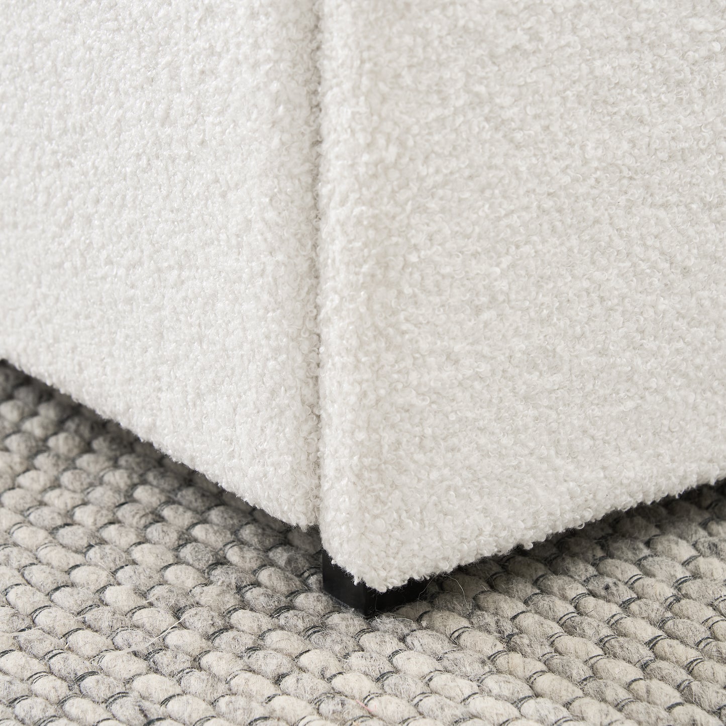 Closeup of a white boucle fabric ottoman corner with black legs on a textured gray rug