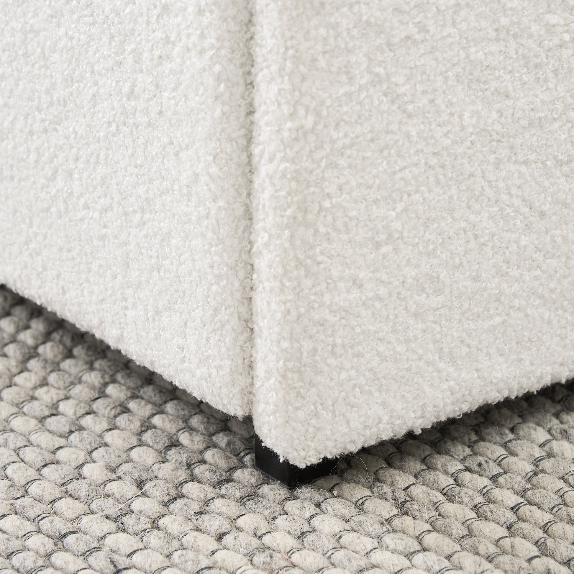 Closeup of a white boucle fabric ottoman corner with black legs on a textured gray rug